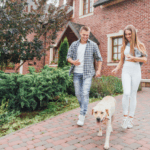 Young homeowner couple takes their dog on a walk and discusses market value vs. replacement cost value of their brick home.